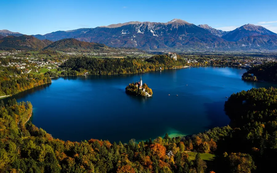 Lake Bled from above in deep blue, the island church in the middle and forested mountains behind