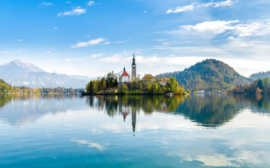 The island church of Lake Bled on its wooded islet, with snow-topped mountains and clouds reflected in the still water