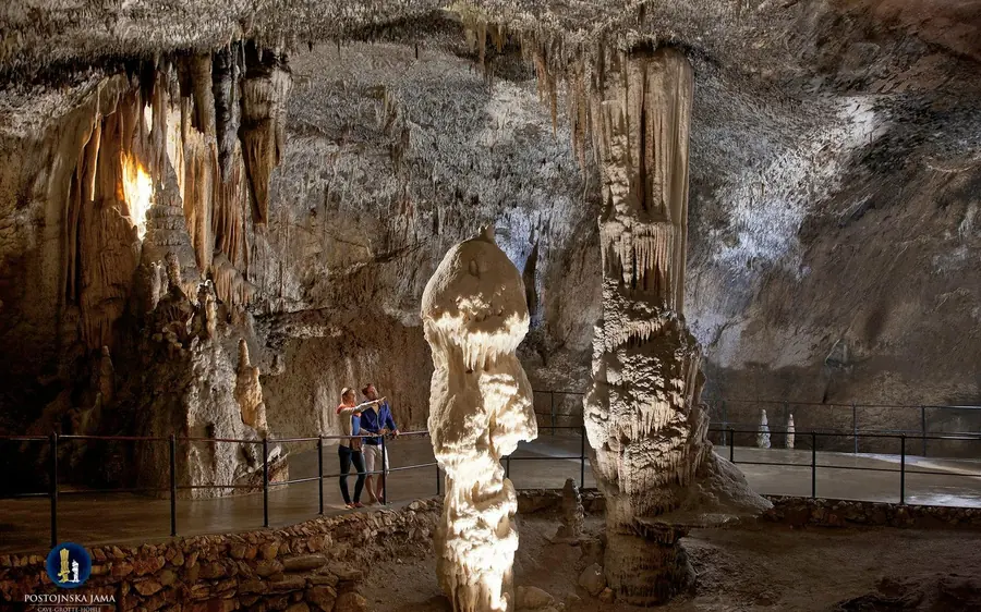 A tall pale stalagmite column in a lit cave chamber, with a railed walkway passing below it
