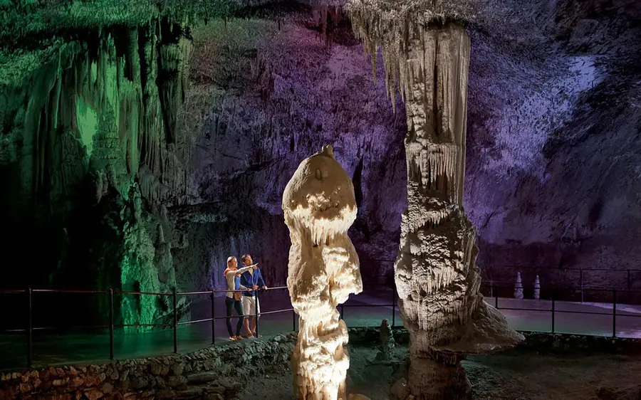 A tall pale cave column lit in green and violet, with two visitors standing at its foot